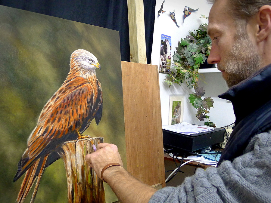 Artist holding a paint brush and painting a red kite that is on a easel