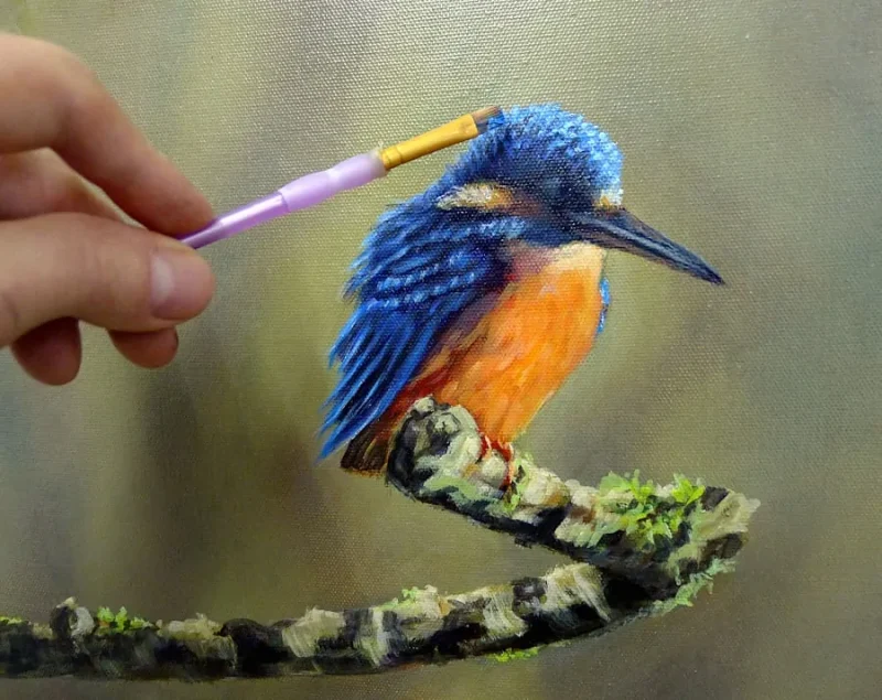 A oil painting of a vibrant kingfisher on an artists easel being painted, with the artists brush touching the canvas.