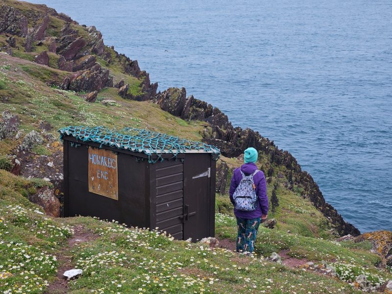 A person with a backpack and hat walking along a grassy cliffside on Skokholm Island, overlooking the ocean, near a small wooden shelter with a sign, during daytime.